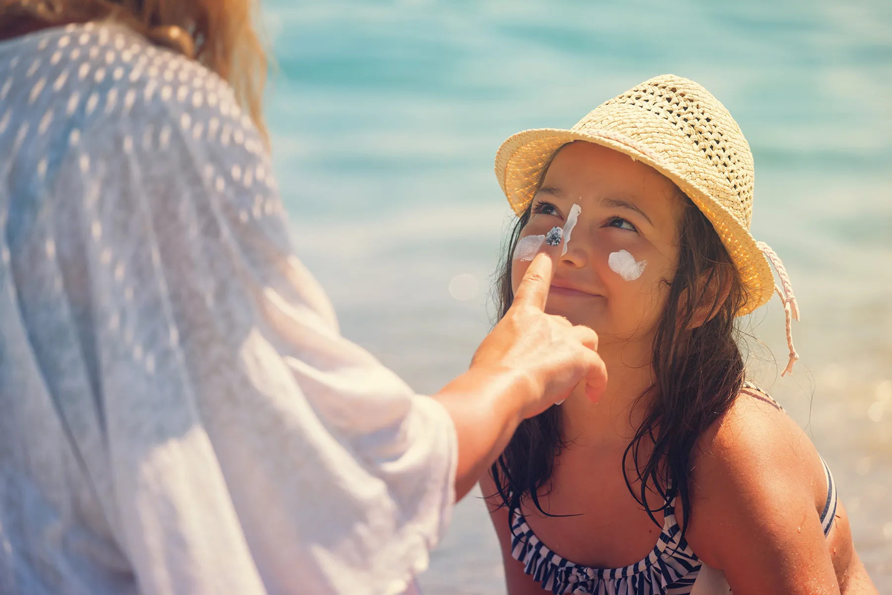 An adult applies white sunscreen to a smiling young girl's face at a sunny beach. The girl wears a straw hat and a striped swimsuit, with the bright blue ocean in the background.