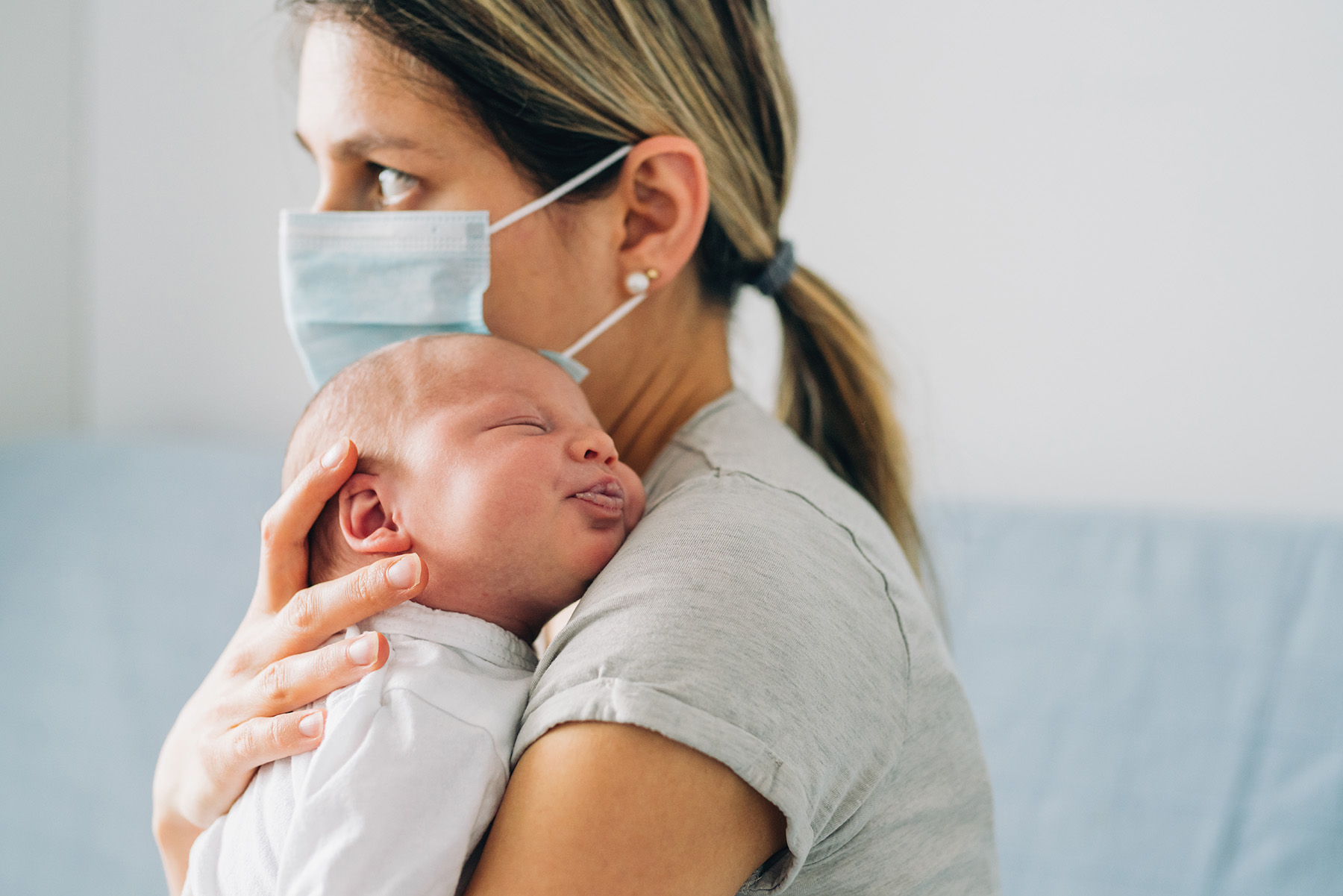 Mother with protective mask holding her newborn baby girl at home.