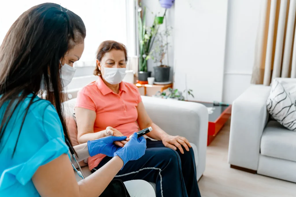 Masked healthcare professional wearing gloves using a glucometer to check an older woman's blood sugar during a home visit.