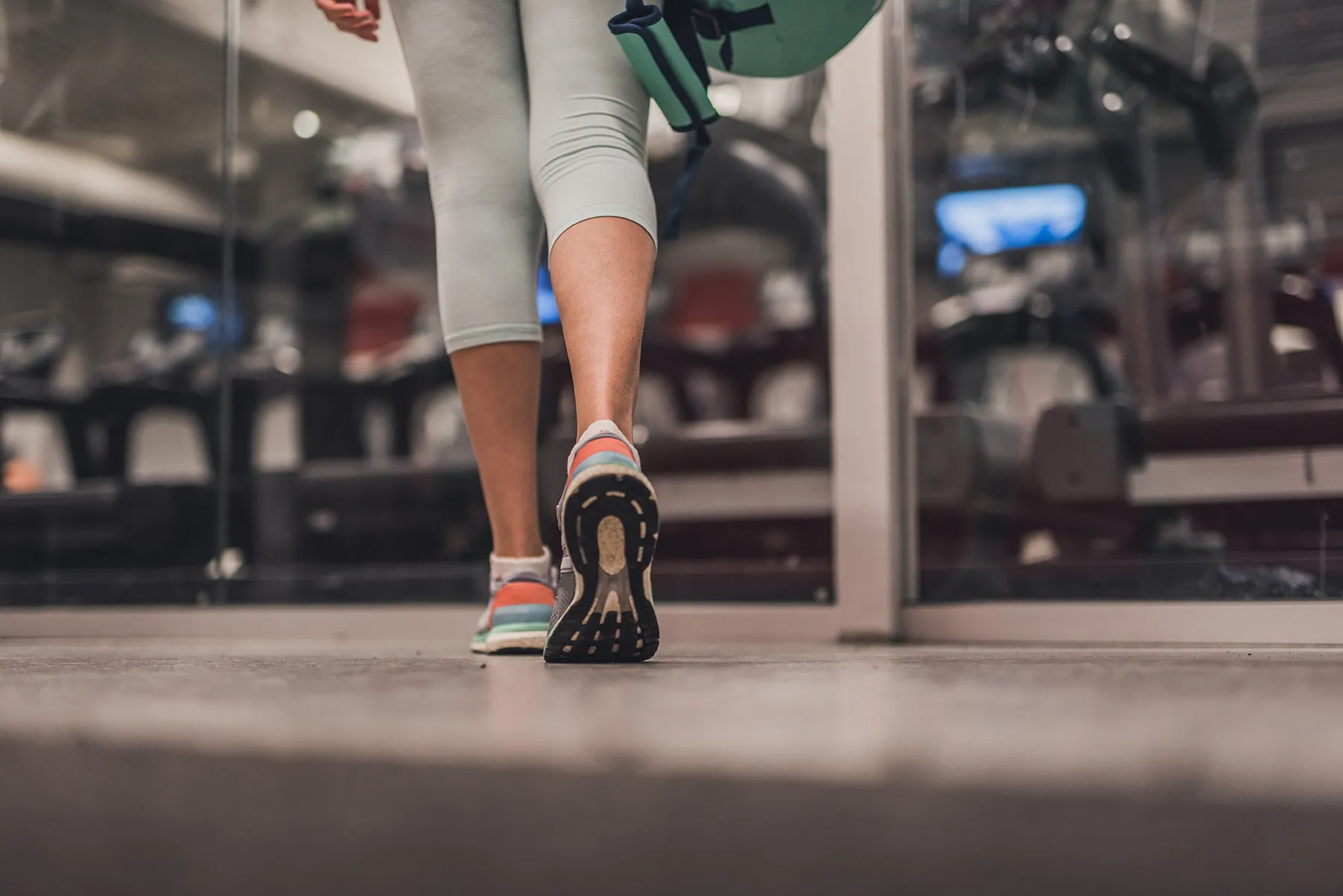 A person's legs, clad in light capri leggings and colorful running shoes, walk away on a gym floor while carrying a green fitness item. Blurred exercise machines and glass walls are visible in the background.