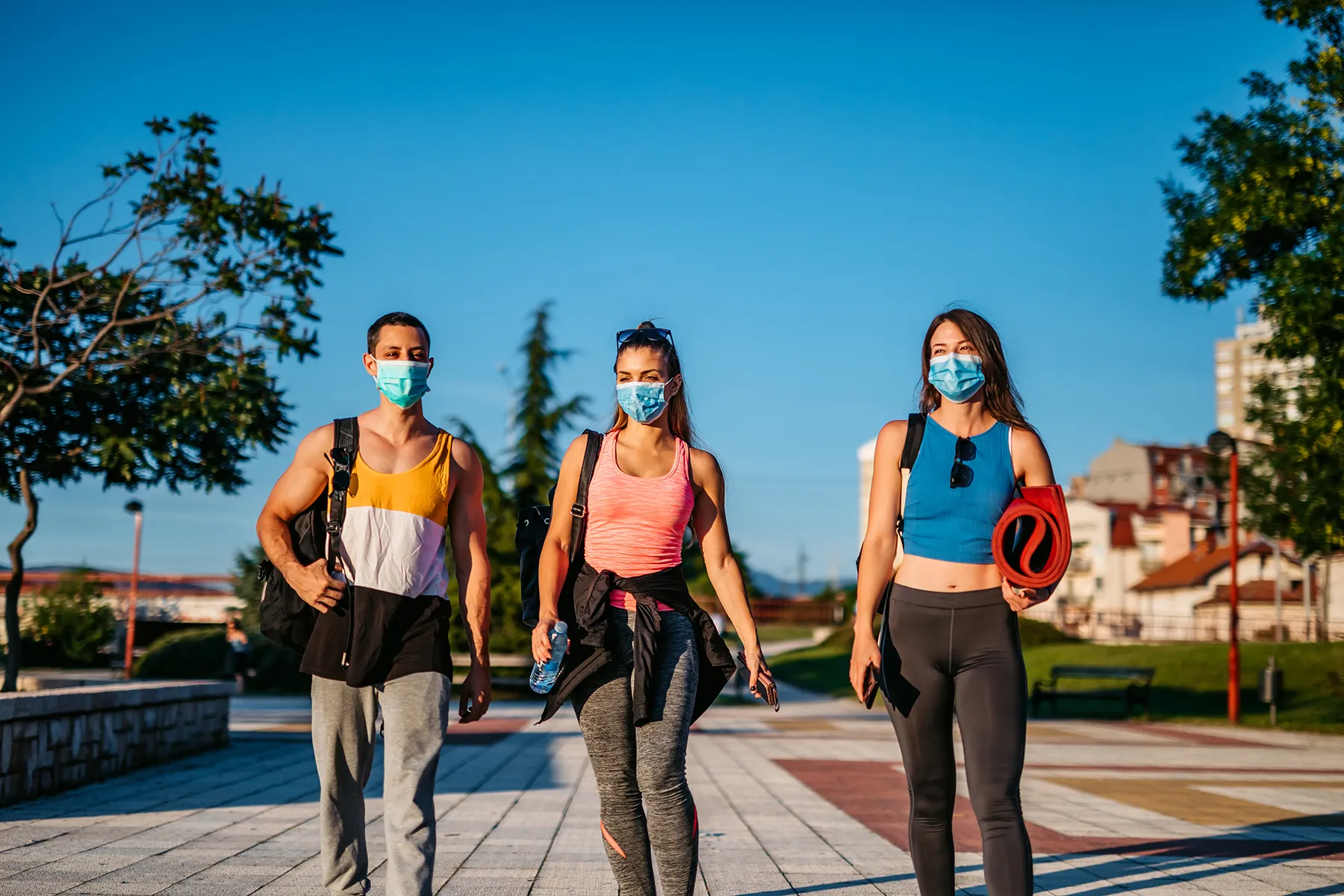 Three friends in face masks and activewear walking outdoors on a sunny day, one holding a red yoga mat.