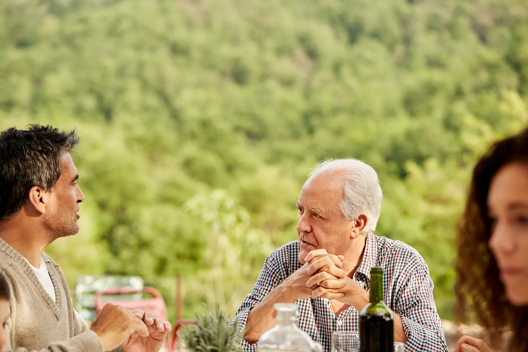 Multi-generational group of people gathered at an outdoor table, featuring an older man with clasped hands and a younger man, engaged in conversation against a lush green hillside background. A wine bottle is on the table.