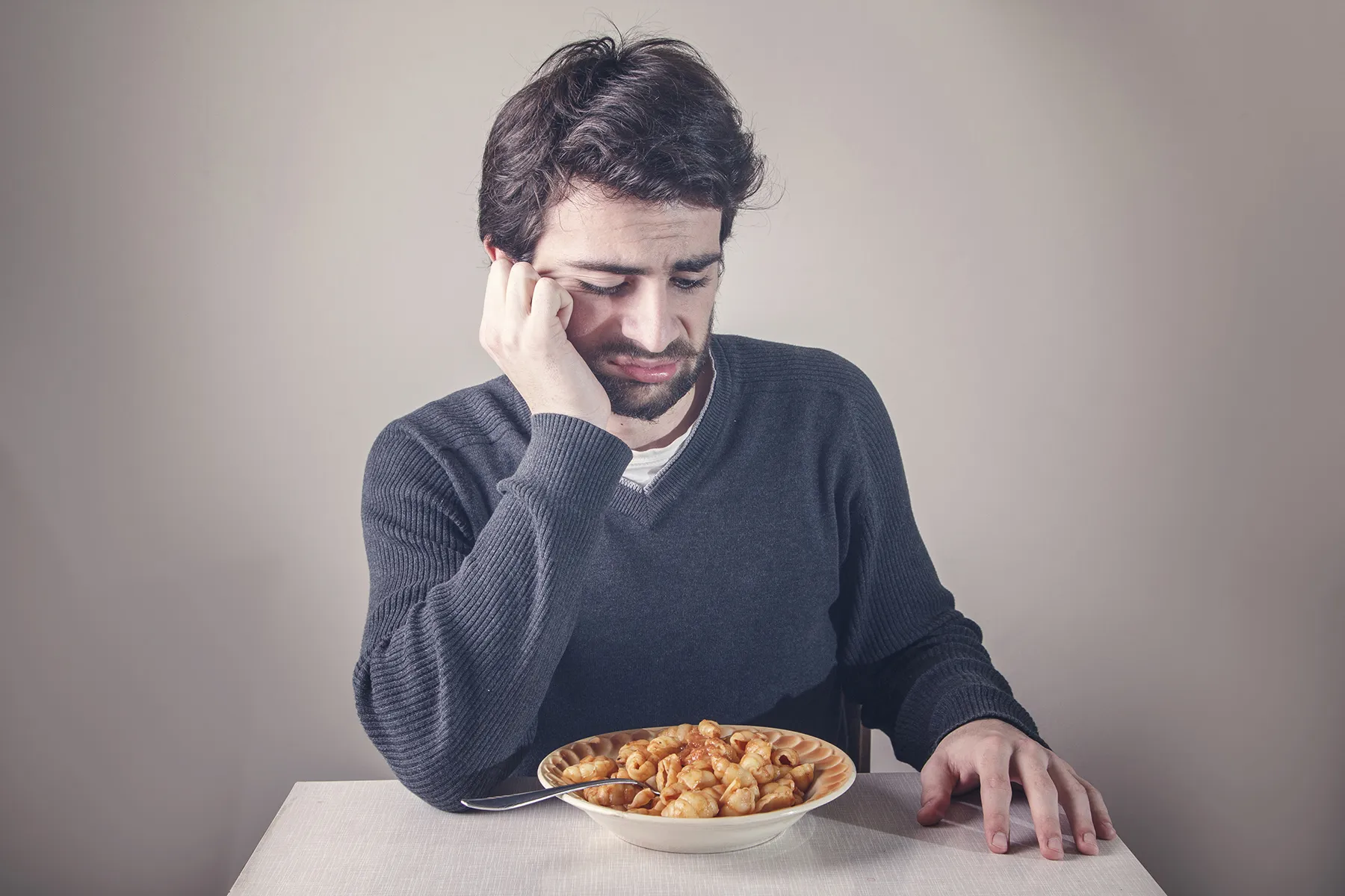 A dejected man with his head resting on his hand, looking down at a bowl of pasta with an expression of disinterest or loss of appetite.