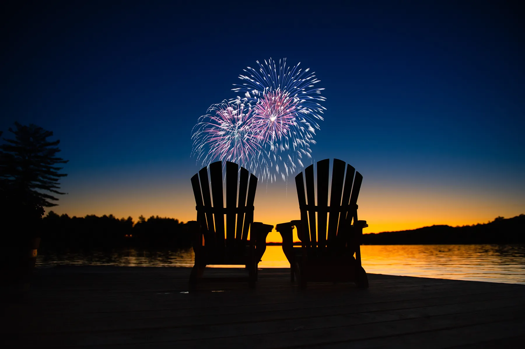 Silhouetted Adirondack chairs on a lake dock at dusk, with colorful fireworks bursting above a sunset horizon.