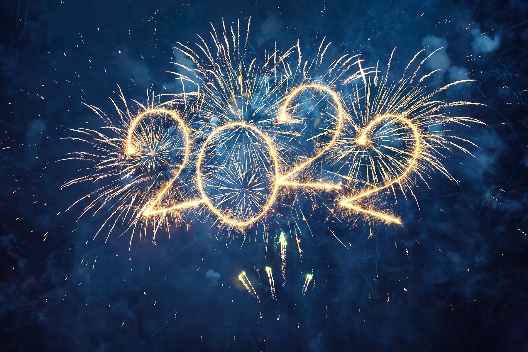 Golden "2022" formed by sparkler trails, set against vibrant fireworks bursting in a dark, smoky night sky.