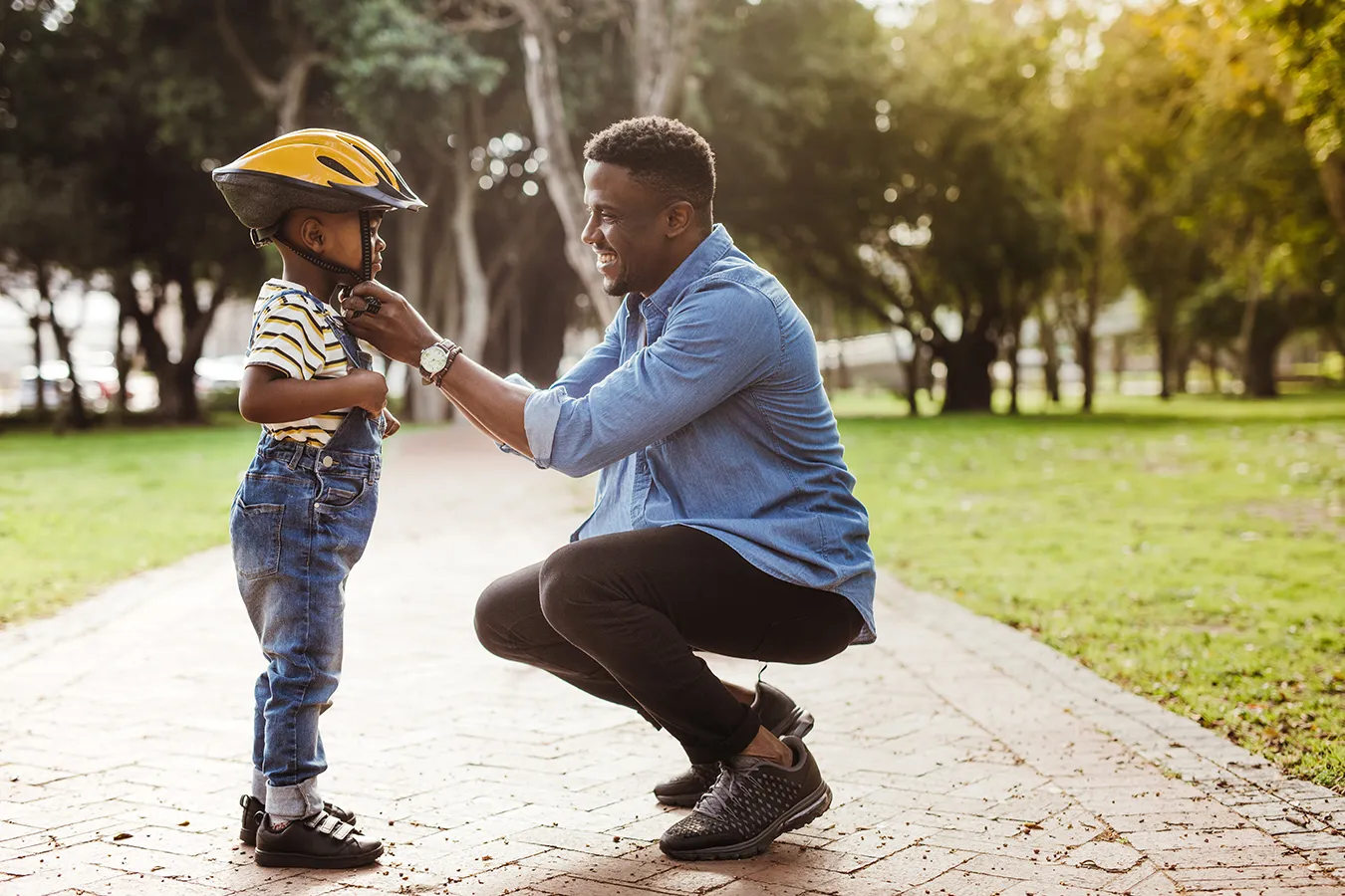 Father helping his young son secure a yellow bike helmet on a park path.