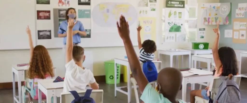 Diverse elementary students actively raise their hands in a bright classroom, led by a masked female teacher at the whiteboard. The classroom is decorated with a world map and educational posters promoting recycling and sustainability.