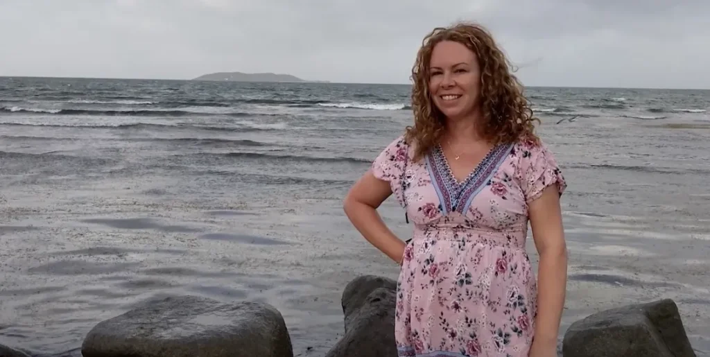 Smiling woman with curly hair in a pink floral dress, standing by choppy grey water with foreground rocks and a distant island under an overcast sky.