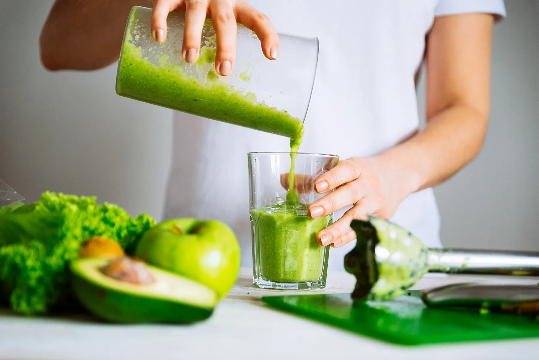Hands pouring a vibrant green smoothie into a glass, surrounded by fresh ingredients like avocado, apple, and lettuce, with an immersion blender on the counter.