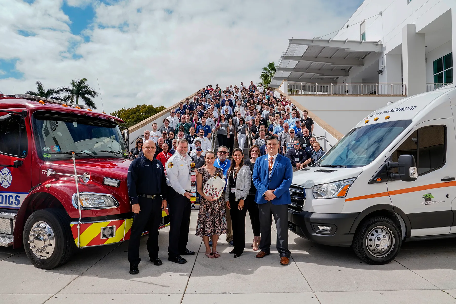 Large group of conference attendees, including first responders and medical personnel, posed on a staircase between a red fire rescue truck and a white medical transport van from The Salah Foundation.