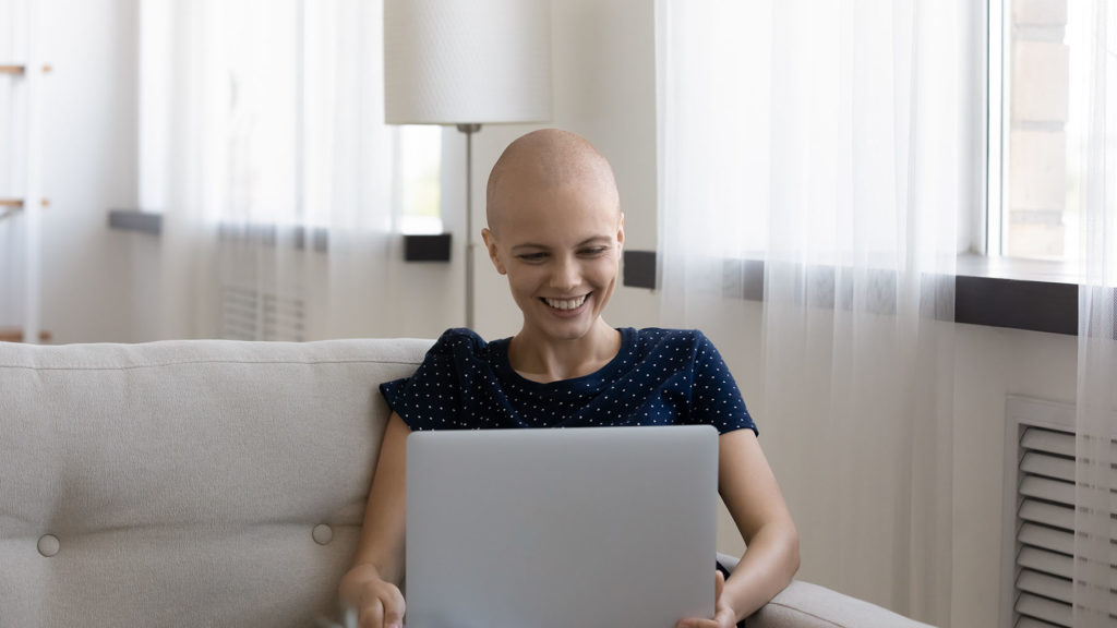 Young cancer patient on couch smiles at laptop