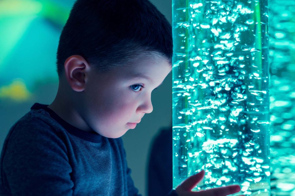 Young boy leans on olored lights bubble tube lamp during therapy session