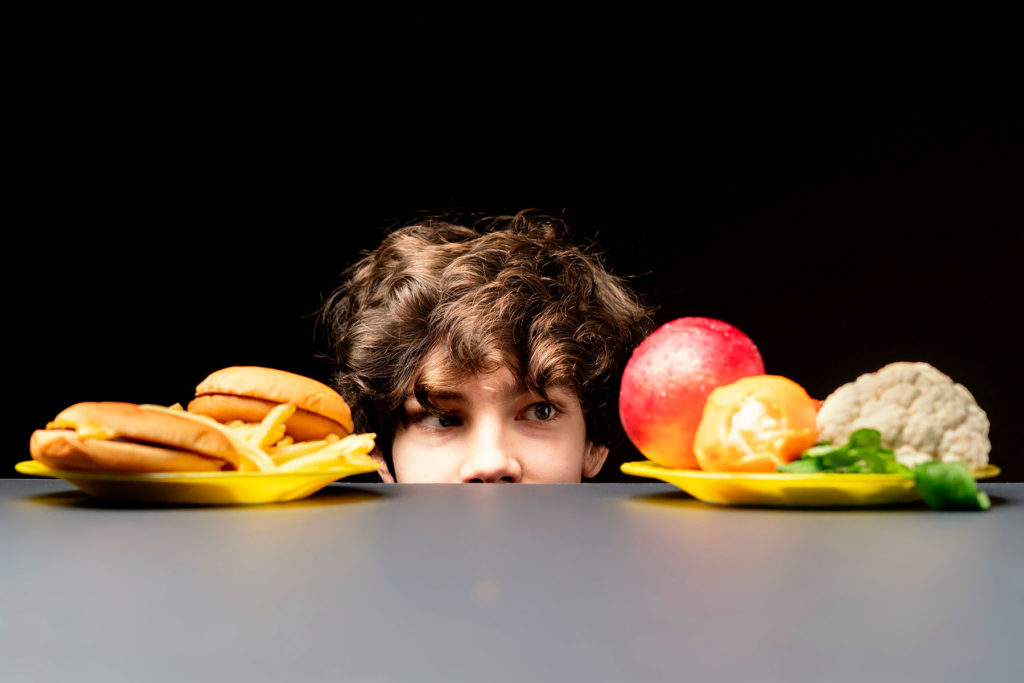 Kid peering out from table with fast food and healthy food choices