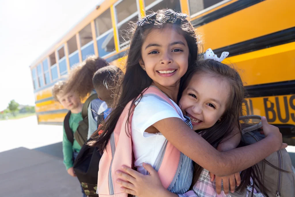 Two young girls smiling and hugging in front of a yellow school bus with other children.