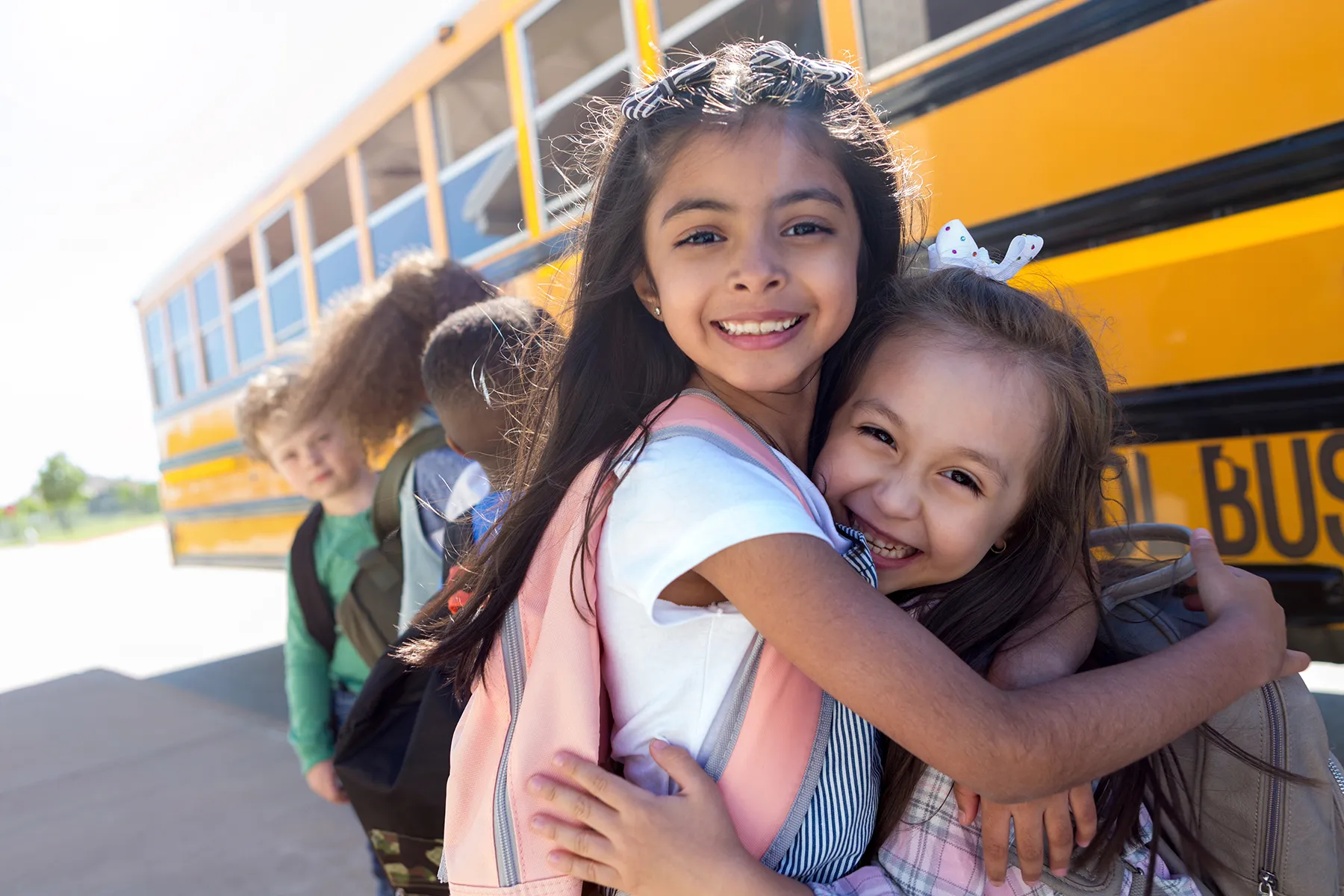 Two young girls smiling and hugging in front of a yellow school bus with other children.