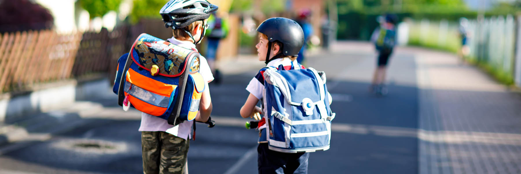Two boys talk while riding scooters and wearing helmets and school backpacks
