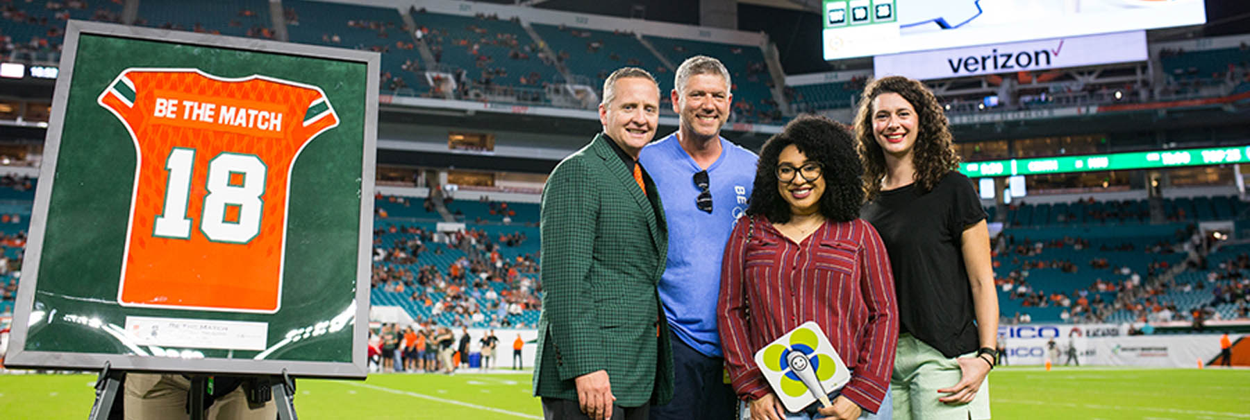 Photo of four adults standing on a football field next to a framed UM Hurricanes football jersey numbered 18 with "Be the Match" printed on it