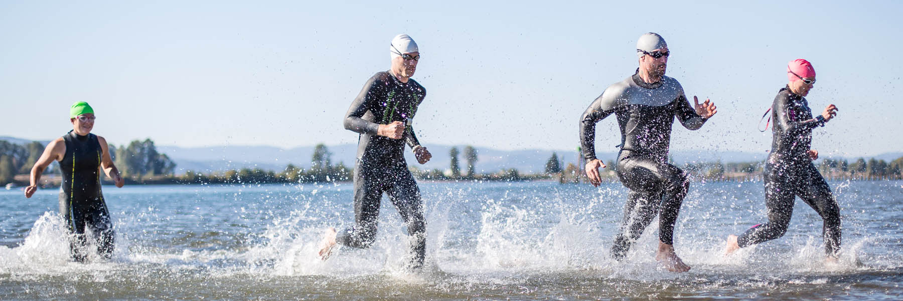 Four athletes wearing wetsuits and swimming goggles running through a body of water