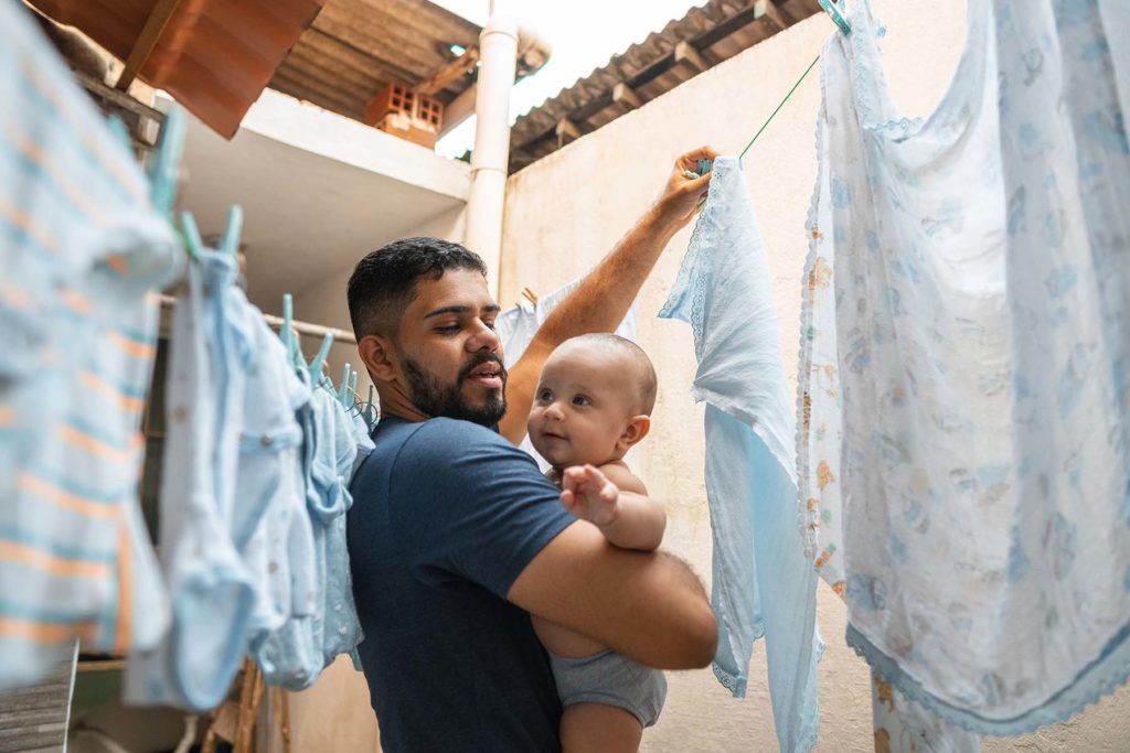 Hispanic dad holds newborn while hanging clothes on a clothesline.