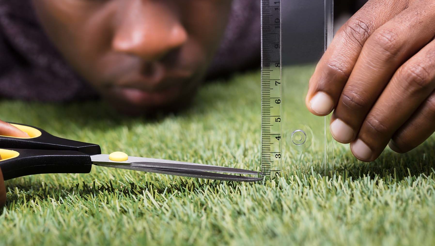 Perfectionist man lies in grass with ruler and scissors to measure grass blades.