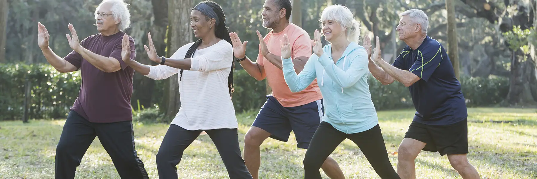 A group of five multi-ethnic seniors taking an exercise class in the park. They are practicing tai chi, standing with their hands raised.