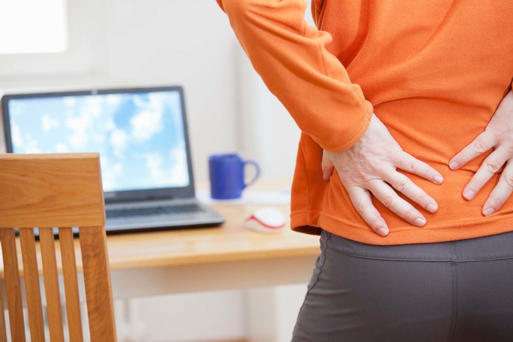 Person in orange shirt holding back in front of computer. Pain from too much sitting.