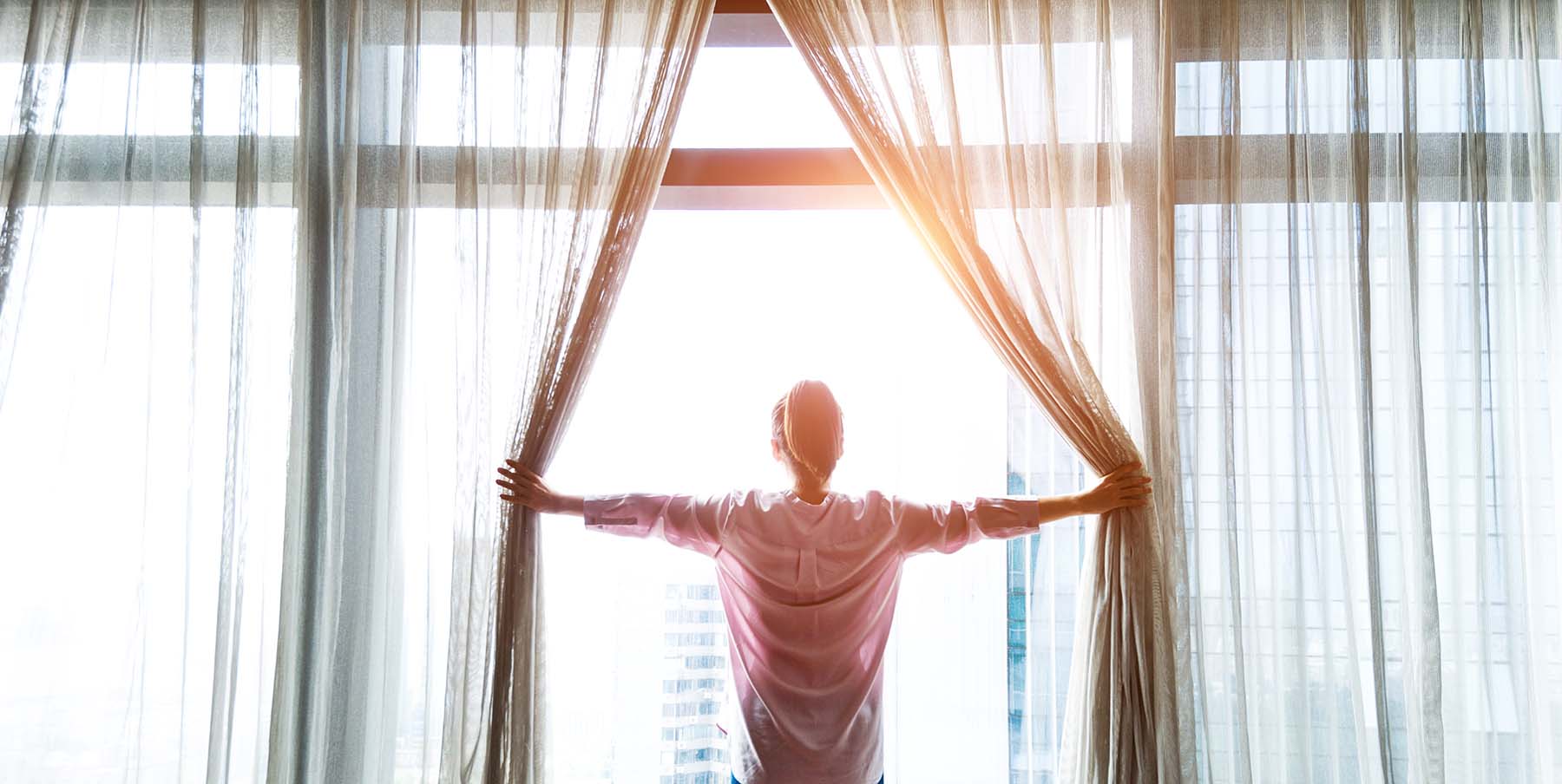 Woman opens curtains to the morning sun overlooks the cityscape.
