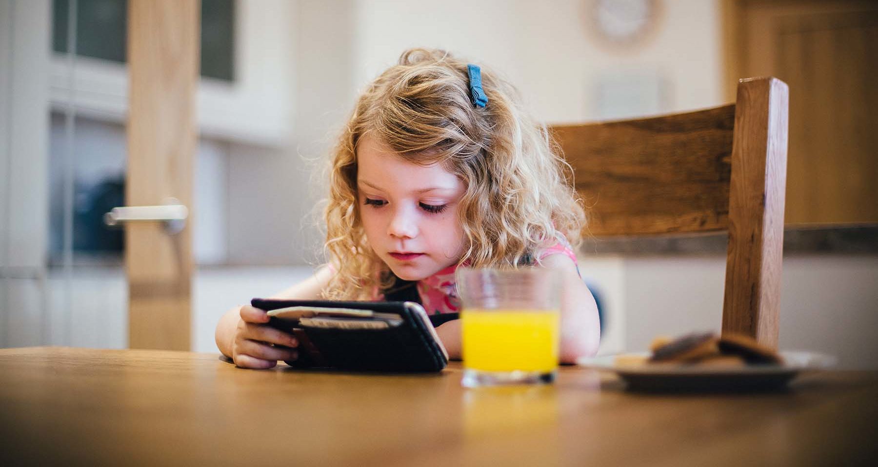 Little blond girl watches smartphone while eating cookies.