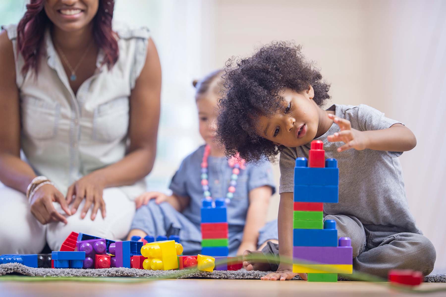 Young Black boy focuses on building blocks with mom and young girl in the background.