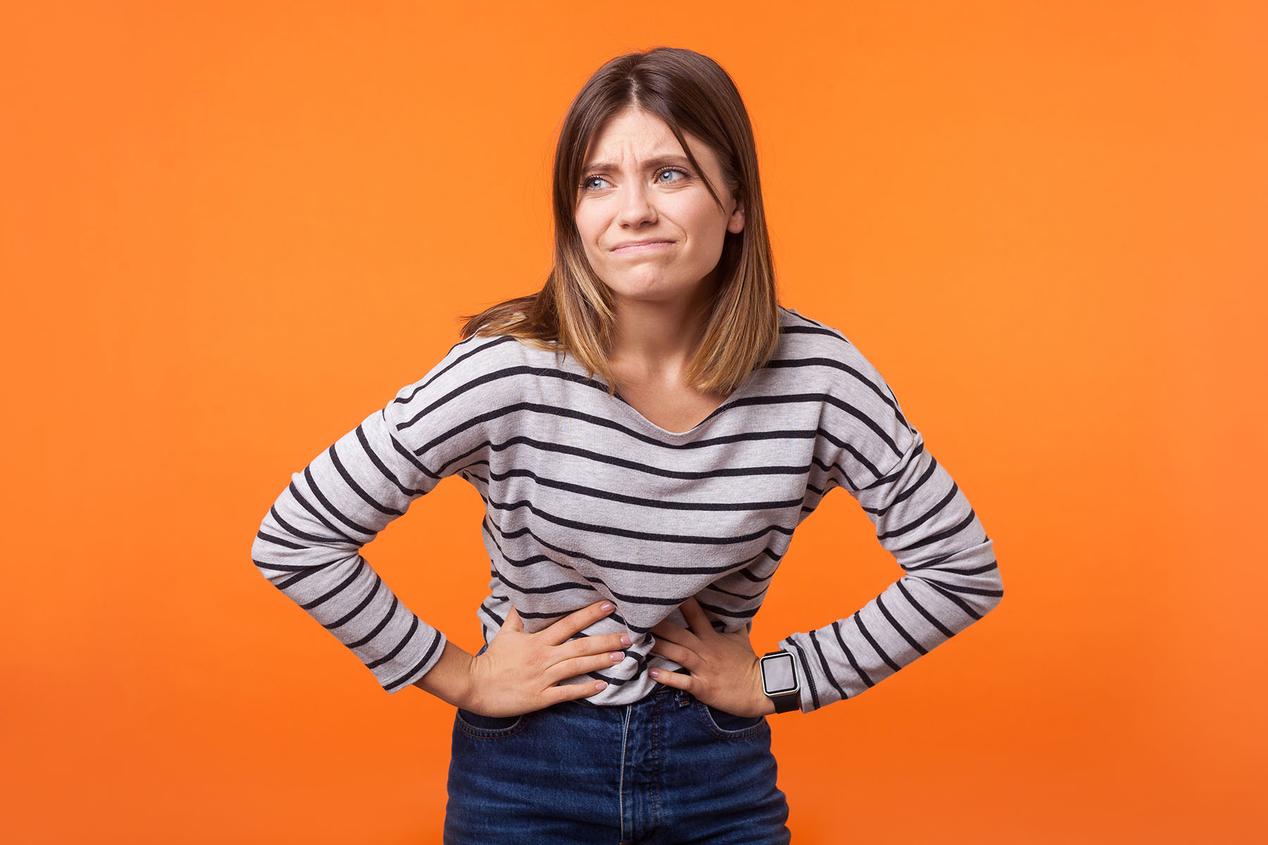 Young woman in striped shirt against orange background holds stomach due to bloating.