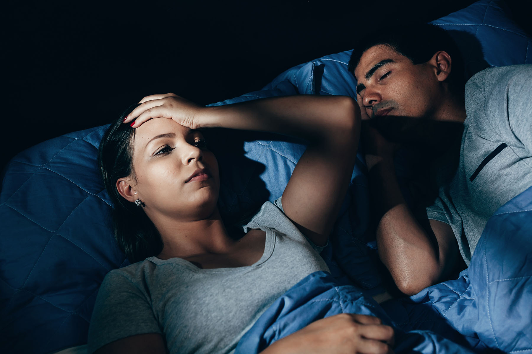 Hispanic couple lies in bed. The man is asleep but the woman has her hand to forehead. Looks stressed.