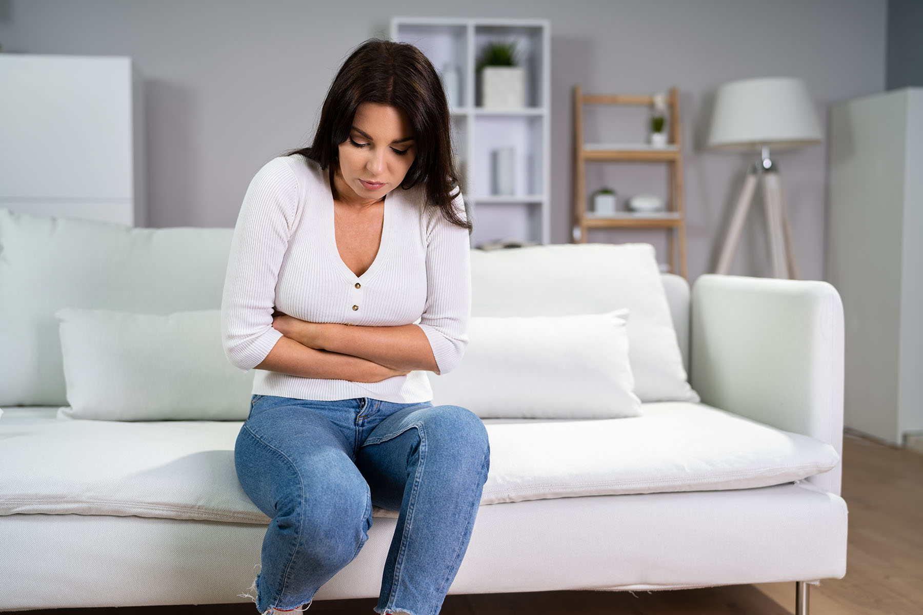 Woman in white shirt sits on couch holding her stomach.