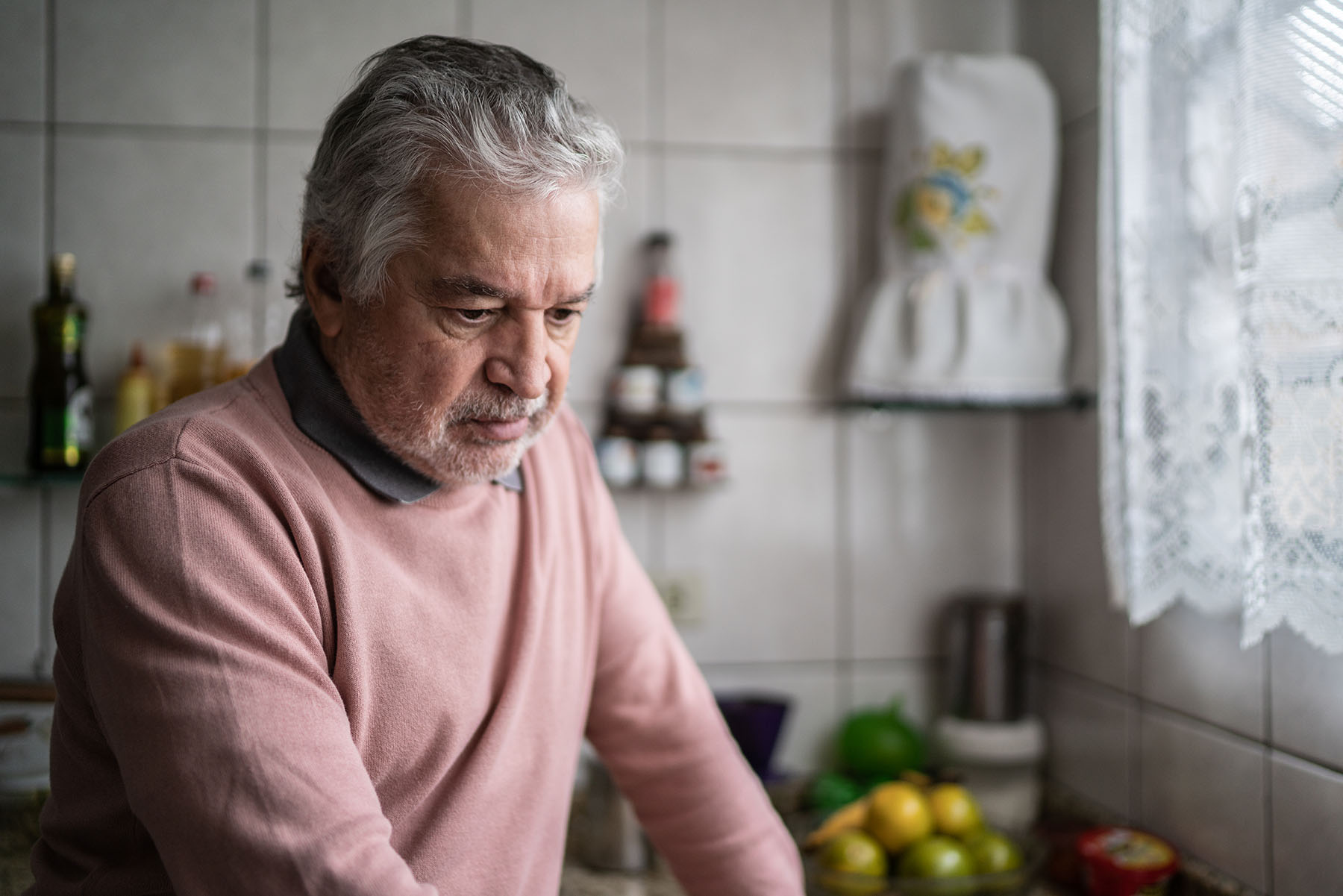 Older Hispanic man leans against kitchen counter, is worried or sad.