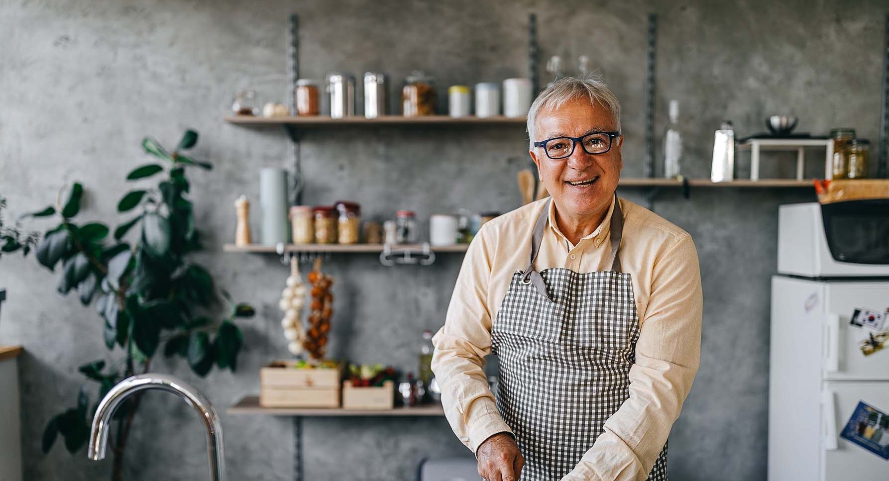 Happy senior man chops vegetables in a minimalistic but cool looking kitchen with gray walls.