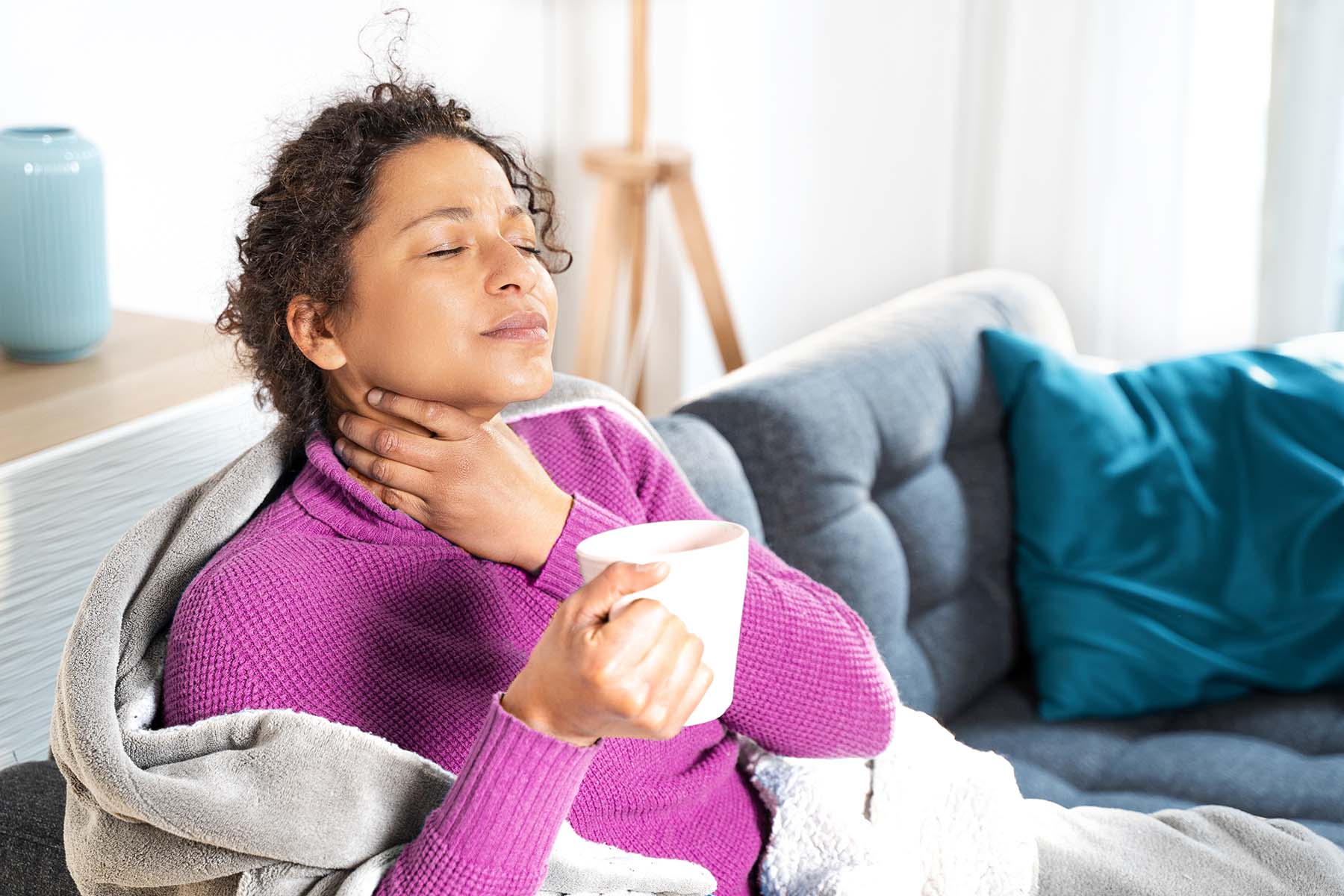 Black woman drinks tea on couch tending to an obviously sore throat.
