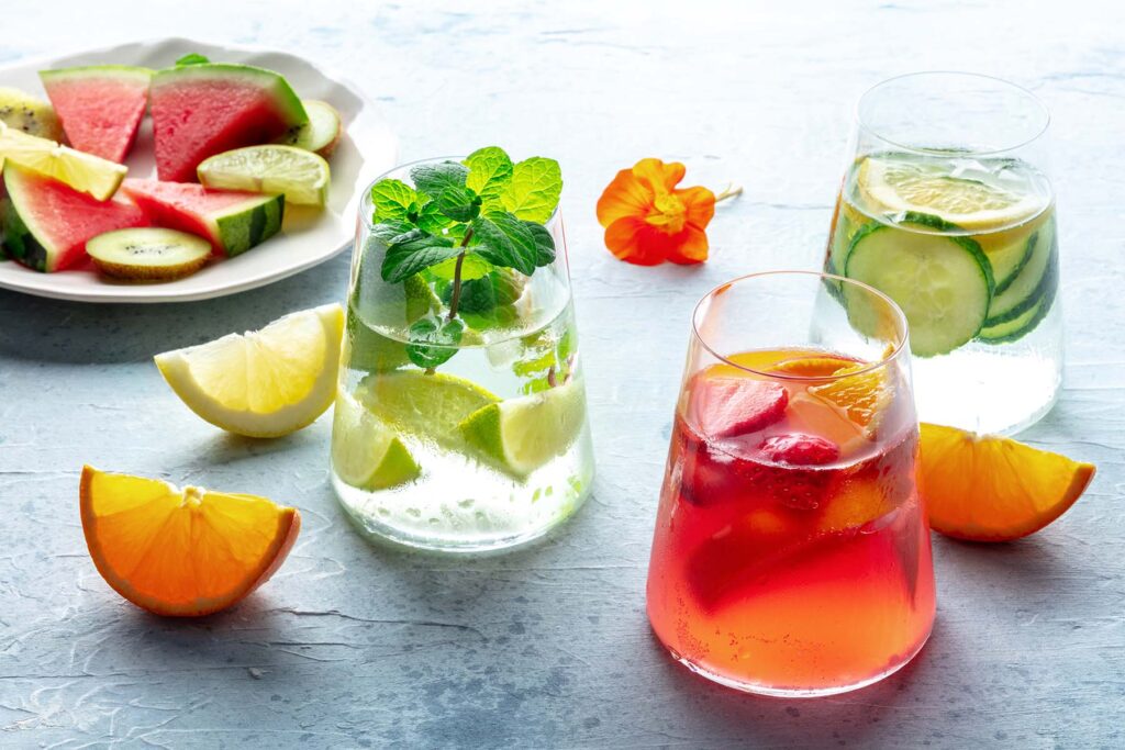 A collection of colorful mocktails on a brightly lit table, along with plates of fruit dsices.