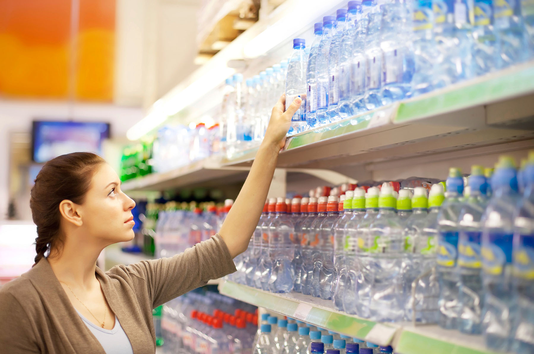 Woman reaches for water bottle on top shelf at grocery store.