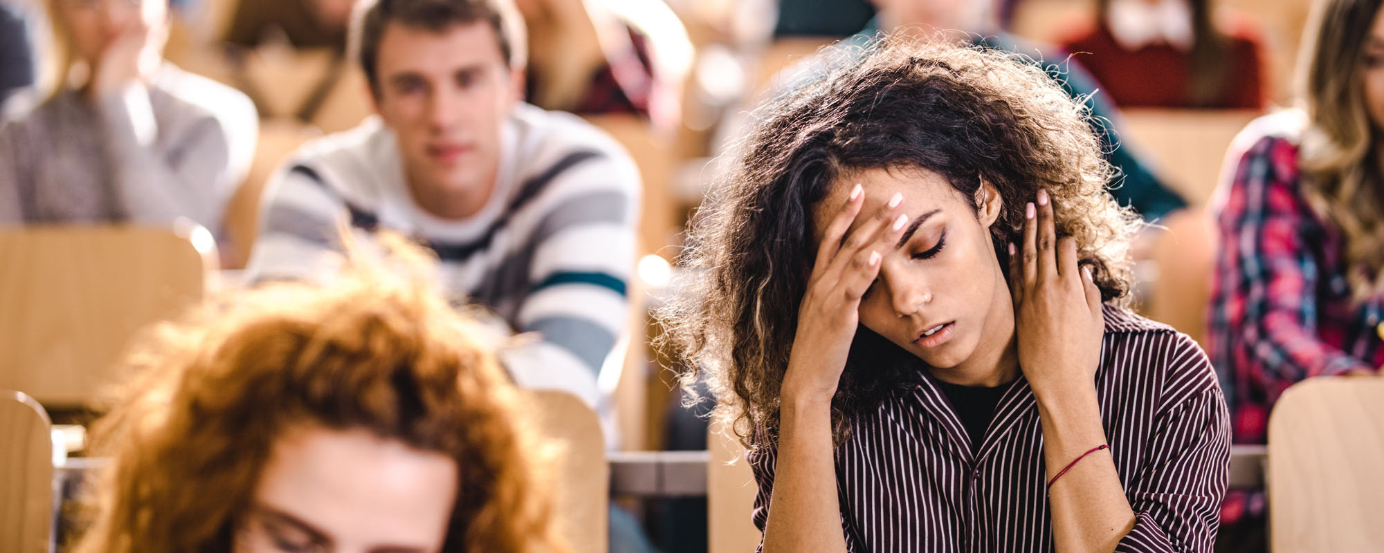 Young adult, college-aged students are sitting in class. Young Black woman is holding her head due to an oncoming migraine headache.