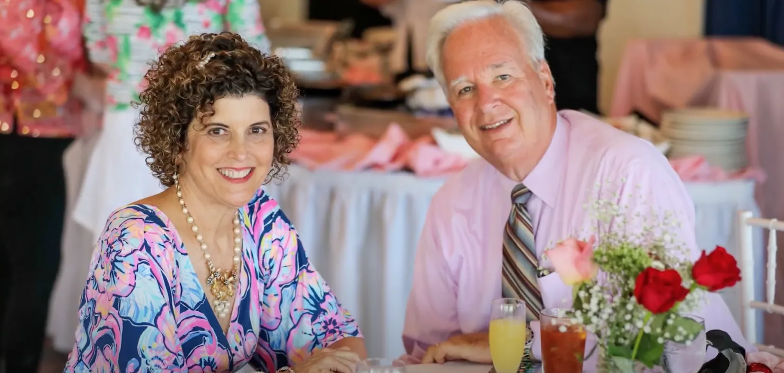 A happy couple smiling at the camera, seated at a table during a celebratory event. The woman wears a brightly patterned dress and a pearl necklace, while the man wears a pink shirt and striped tie. Pink decor and flowers are visible in the background.