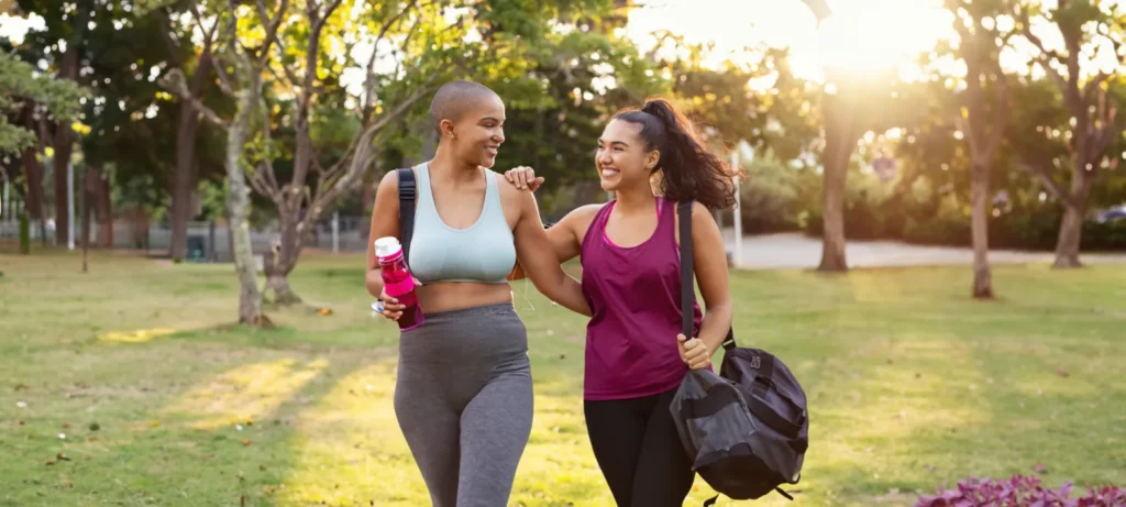 Two smiling women in athletic wear walking together through a sunny park. One woman carries a pink water bottle, and the other has a black gym bag.