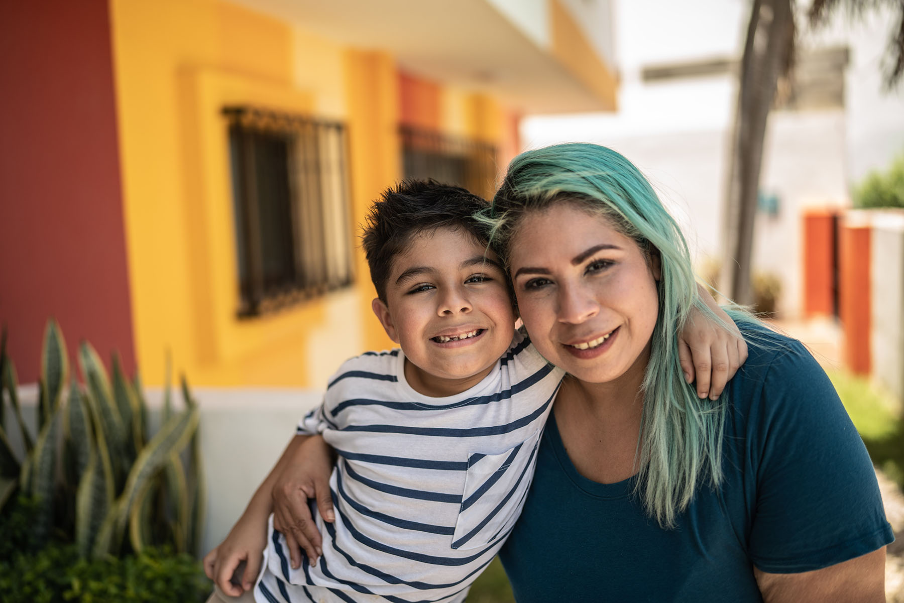 Hispanic mother with hair dyed green smiles with her son outside a house.