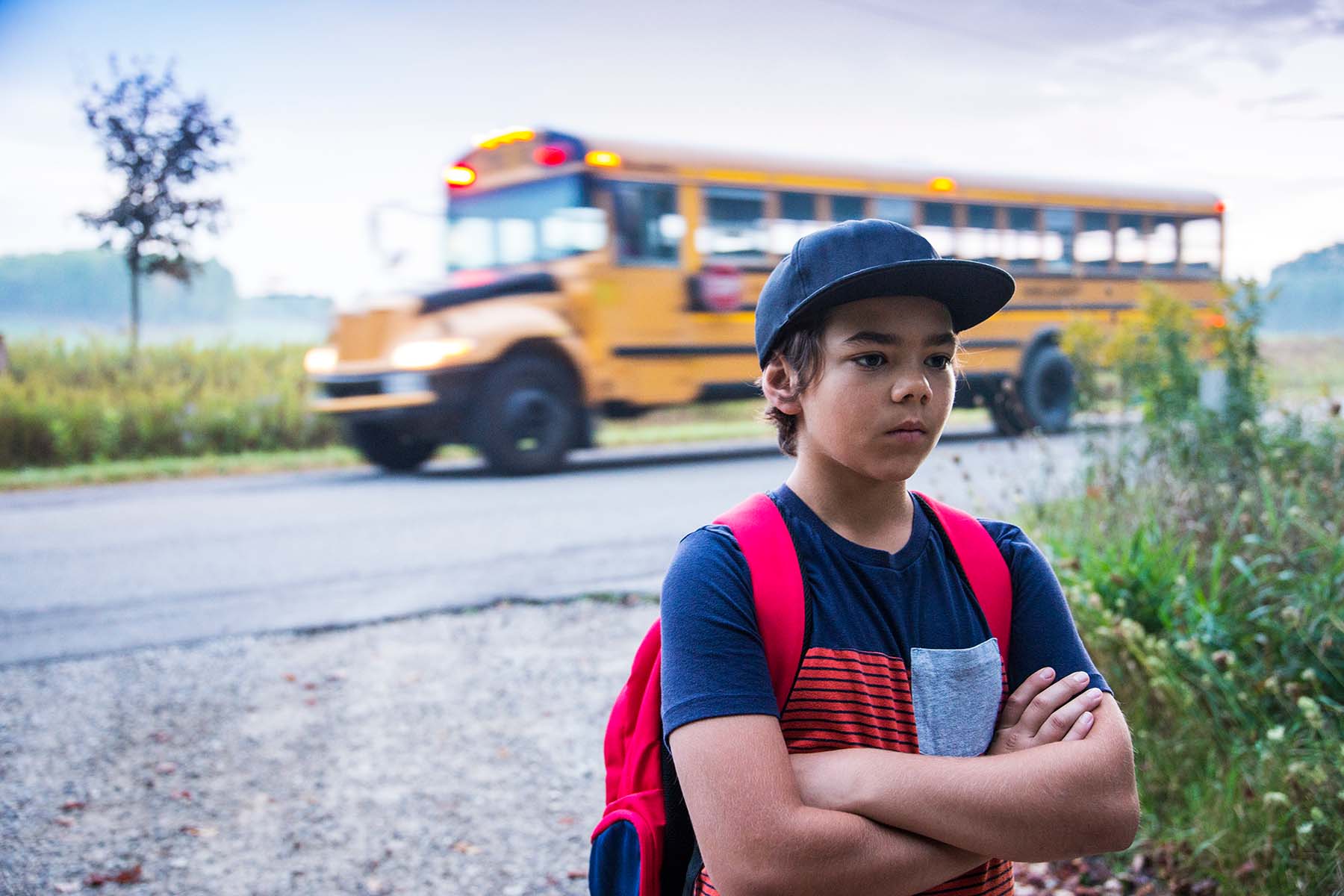 Grumpy young boy stands with his back to school bus, pouting.