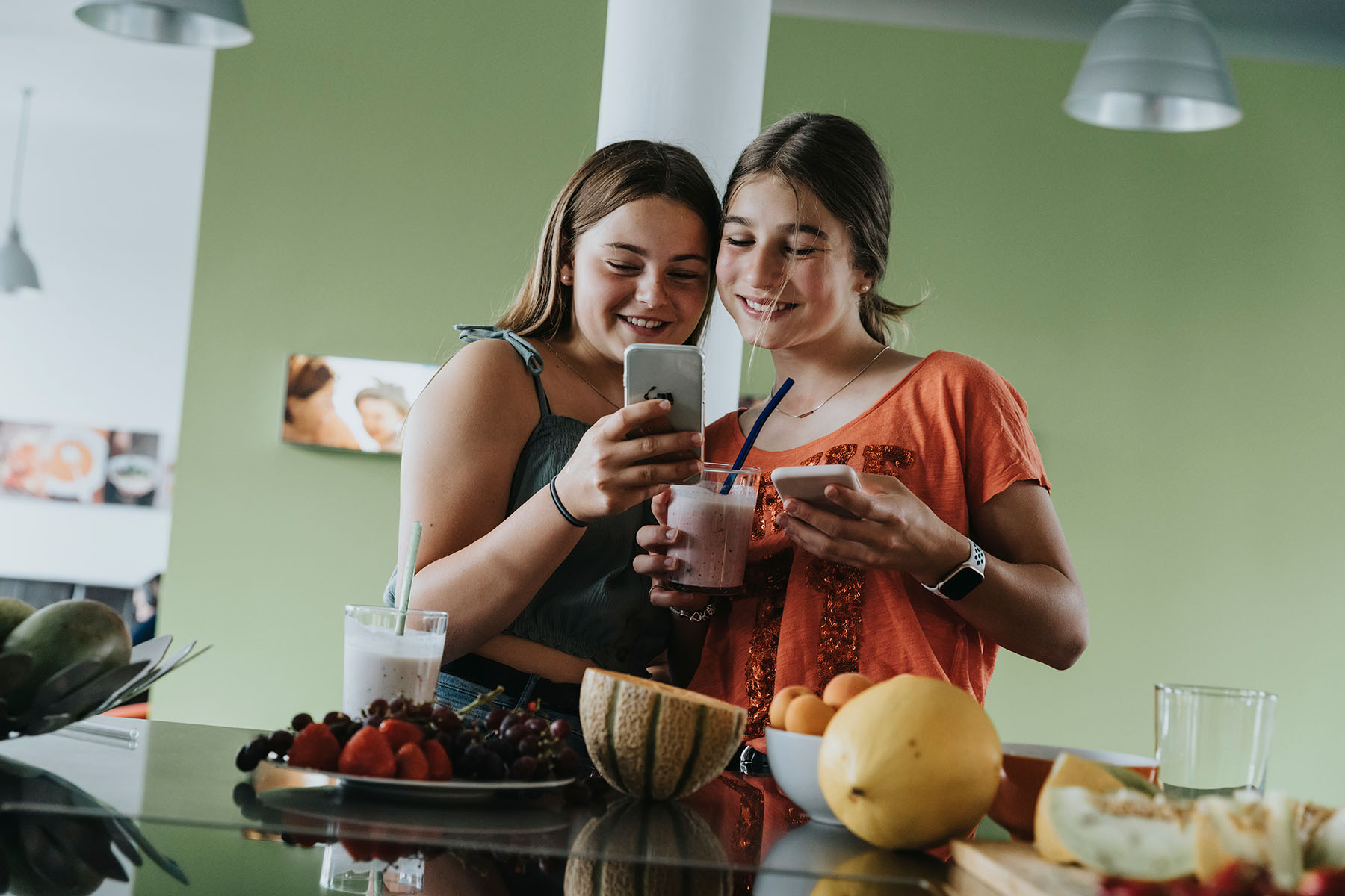 Two girls smiling as the enjoy homemade fruit smoothies and share cell phone photos.