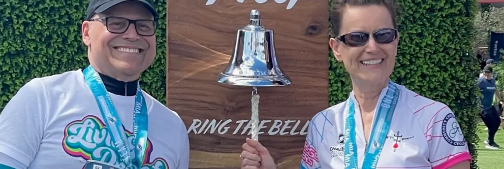 Smiling man and woman in athletic wear, displaying event medals, celebrate an endurance or charity challenge completion by ringing a large silver bell. The bell is mounted on a wooden "RING THE BELL" sign in an outdoor setting with green foliage.