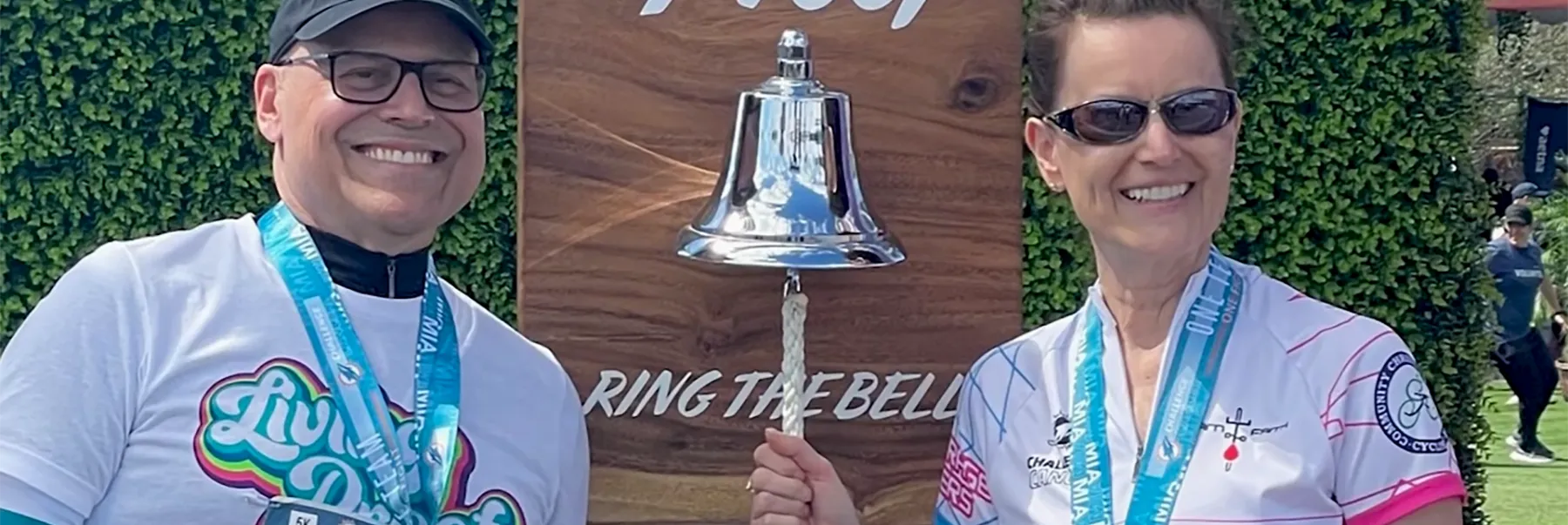 Smiling man and woman in athletic wear, displaying event medals, celebrate an endurance or charity challenge completion by ringing a large silver bell. The bell is mounted on a wooden "RING THE BELL" sign in an outdoor setting with green foliage.