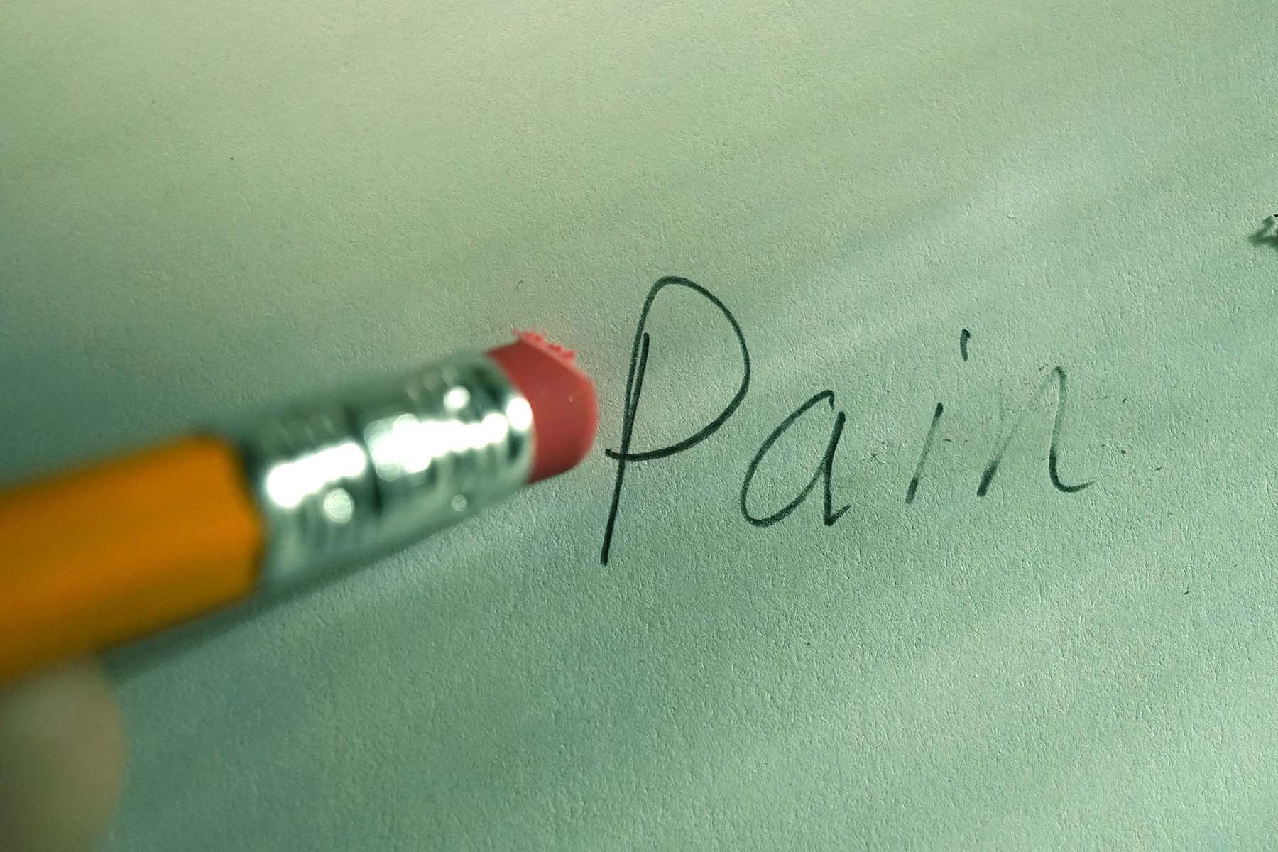 The butt of a pencil is shown erasing the word "Pain," which was written on paper.