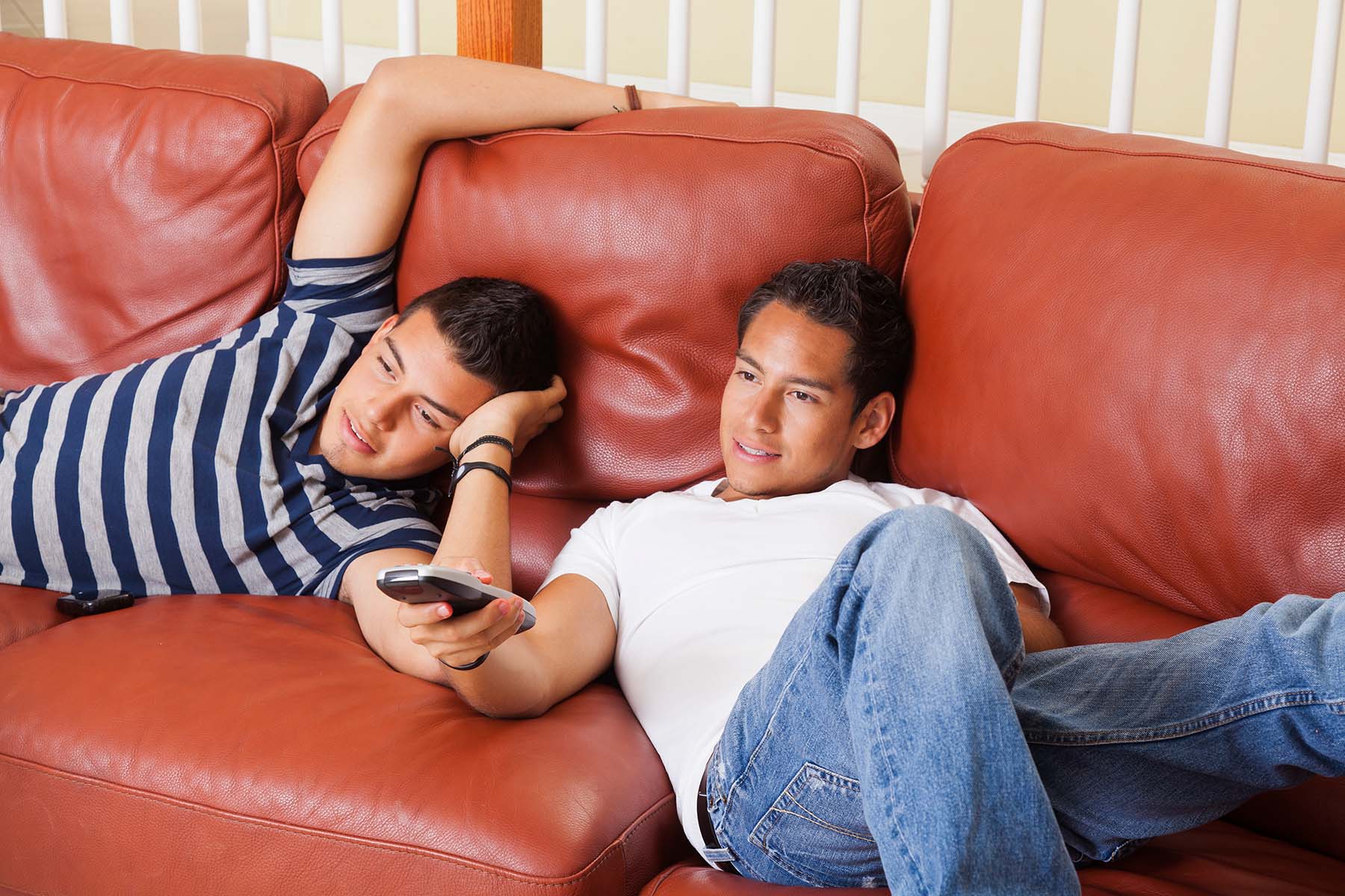 Two Hispanic teen boys flop on a leather couch to watch tv.