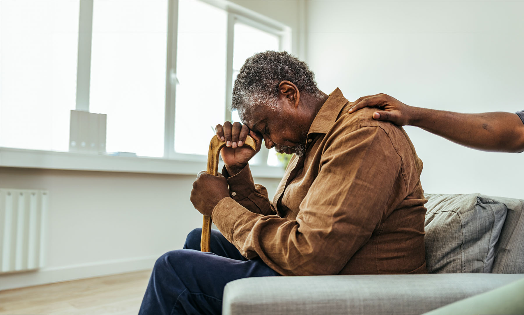 Black elderly man with cognitive decline sits on white couch with his head in his hand because he is stressed. There is a hand on his shoulder trying to comfort him.