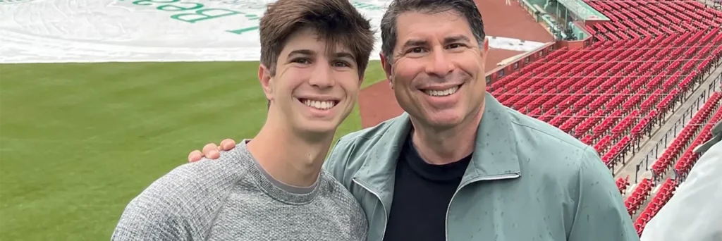 Smiling father and son posing at Fenway Park, with a green baseball field and empty red stadium seats in the background.