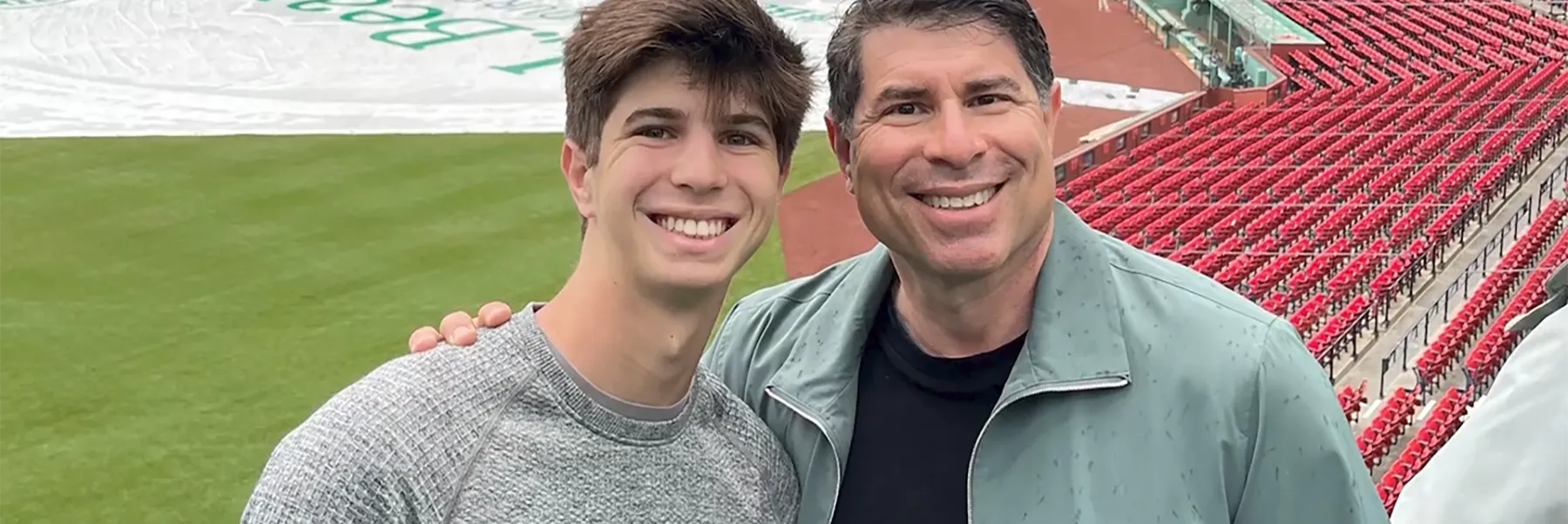 Smiling father and son posing at Fenway Park, with a green baseball field and empty red stadium seats in the background.